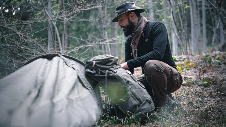 Ein Mann in Outdoor-Kleidung kniet neben Campingausrüstung in einem Waldgebiet.