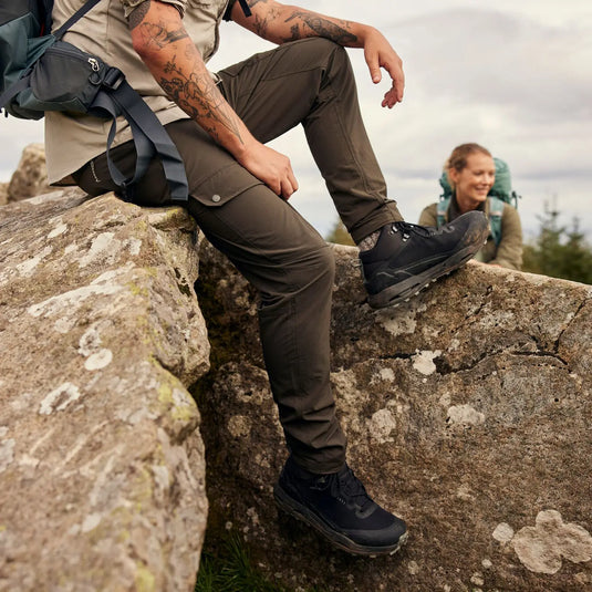 Zwei Wanderer machen auf einem felsigen Ausläufer eine Pause, wobei der Fokus auf den Stiefeln einer Person liegt, die Craghoppers NosiLife Adventure Hose III für Herren bieten.