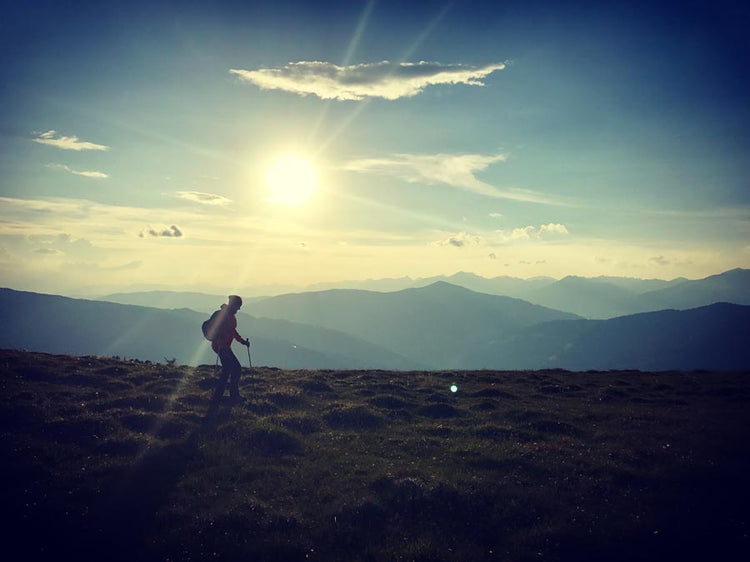 Ein Wanderer erklimmt bei Sonnenuntergang einen grasbewachsenen Hügel vor der Kulisse von Bergketten.