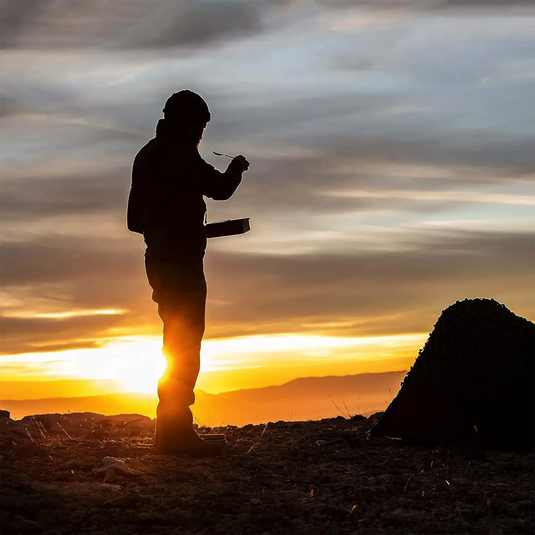 Silhouette einer Person, die bei Sonnenuntergang in der Nähe eines Campingplatzes isst und ein Esbit® Trockenbrennstoff-Kochset 1100 ml für ein Outdoor-Abenteuer verwendet.