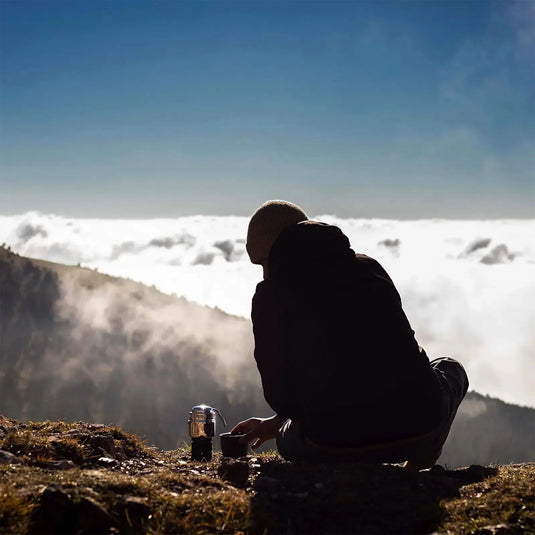 Eine Person sitzt auf einem Berghang und genießt die Aussicht über den Wolken, mit einer Esbit® Reise-Kaffeemaschine an ihrer Seite.