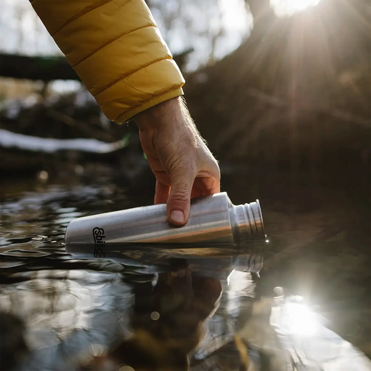 Eine Person in einer gelben Jacke füllt eine Esbit® Trinkflasche Sculptor 1000ml Edelstahl mit 1000ml Wasser aus einem Bach, während Sonnenlicht durch die Bäume scheint.