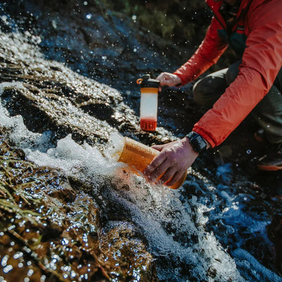 Eine Person entnimmt eine Wasserprobe aus einem Bach, um sie mit dem Grayl Geopress Wasserfilter 710 ml von Grayl® auf Krankheitserreger zu testen.