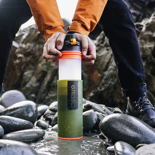 Person mit einer Grayl® Geopress Wasserfilterflasche steht auf nassen Flusssteinen und sichert sich damit sauberes Trinkwasser.