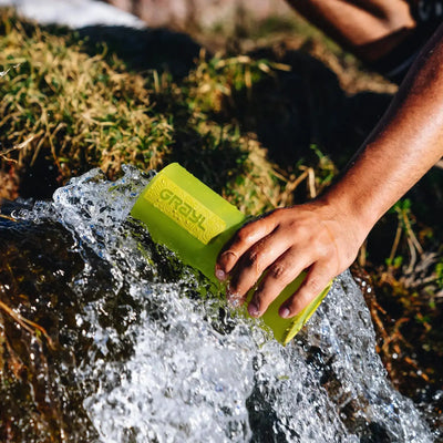 Eine Hand füllt eine grüne Wasserflasche mit Grayl® Ultrapress Wasserfilter 500 ml aus einem fließenden Bach nach.