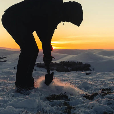 Eine Person gräbt bei Sonnenuntergang mit einem HIGHLANDER® Doppel-Faltspaten im Schnee.