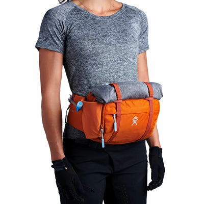 Eine Person, die ein graues T-Shirt, schwarze Handschuhe und einen orangefarbenen Hydro Flask® 5 L Down Shift™ Hydration Hip Pack trägt, an dem eine zusammengerollte Decke befestigt ist.