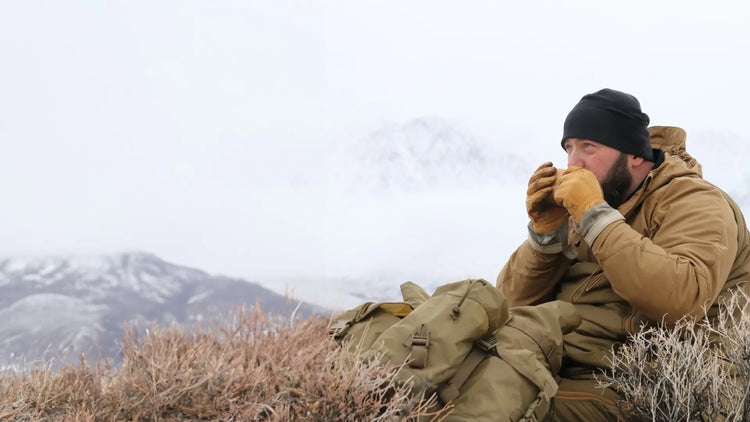 Eine Person sitzt in einer kalten Berglandschaft und wärmt die Hände, während sie ein warmes Getränk in der Hand hält.