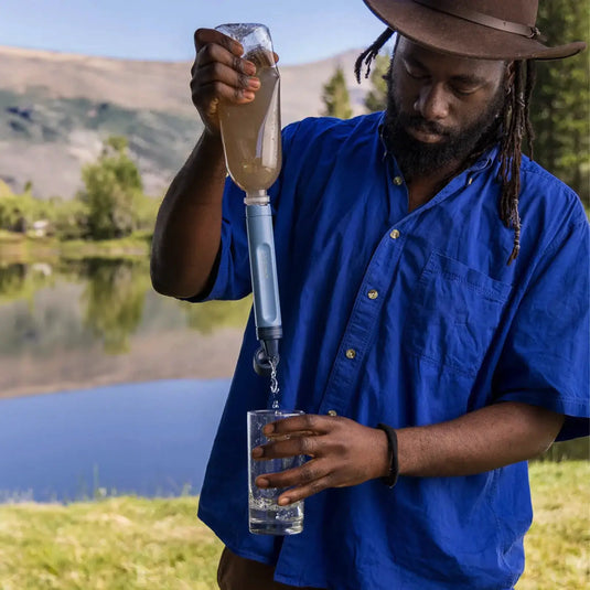 Mann mit Hut füllt mit einem LifeStraw® Peak Series-Strohhalm ein Glas an einem See.
