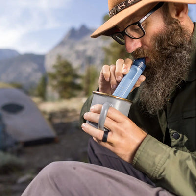 Eine bärtige Person, die aus einem tragbaren LifeStraw® Peak Series Straw-Becher mit Strohhalm trinkt, sitzt draußen in der Nähe eines Zeltes mit Bergen im Hintergrund.