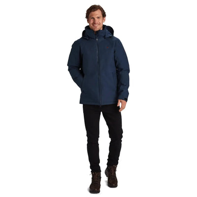 Ein Mann in einer blauen Y by Nordisk® Marton - Herren Hardshell Daunenjacke, schwarzen Hosen und braunen Stiefeln steht mit den Händen an den Seiten und lächelt.