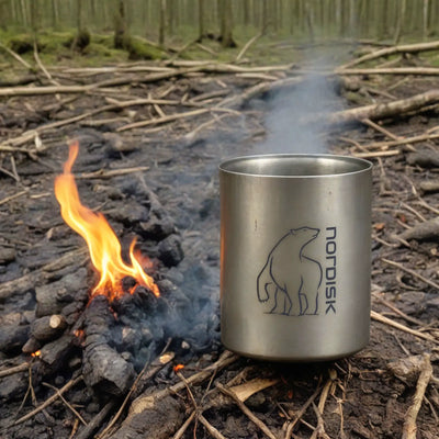 Ein Nordisk®-Titanbecher mit Markenlogo neben einem kleinen Lagerfeuer auf einem Waldboden.