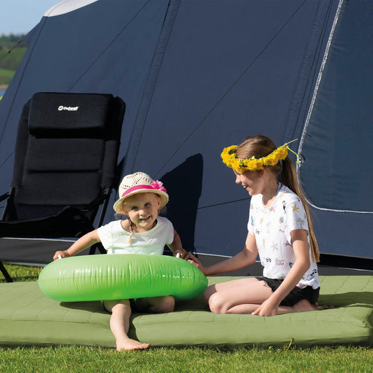 Zwei Kinder mit Blumenkränzen sitzen an einem sonnigen Tag in der Nähe eines Zeltes, während ein Kind ein grünes aufblasbares Outwell® Dreamland in der Hand hält.