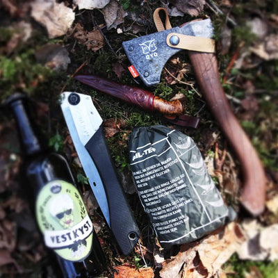 Outdoor-Überlebensausrüstung, darunter eine Axt, ein Messer und Notrationen, mit einer Flasche Bier und einer ØYO Tursag Klappsäge 22 cm auf einem Waldboden.