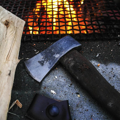 Ein ØYO Solide Sportaxt mit Eschenholzgriff, der auf einer Oberfläche ruht, mit einem Lagerfeuer im Hintergrund.