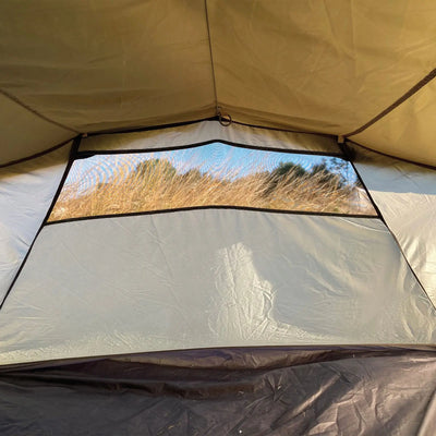 Ein Blick auf hohes Gras durch das abgeschirmte Fenster eines Robens® 4-Personen Zelt Voyager Versa 4 Zeltes, beleuchtet von Sonnenlicht.