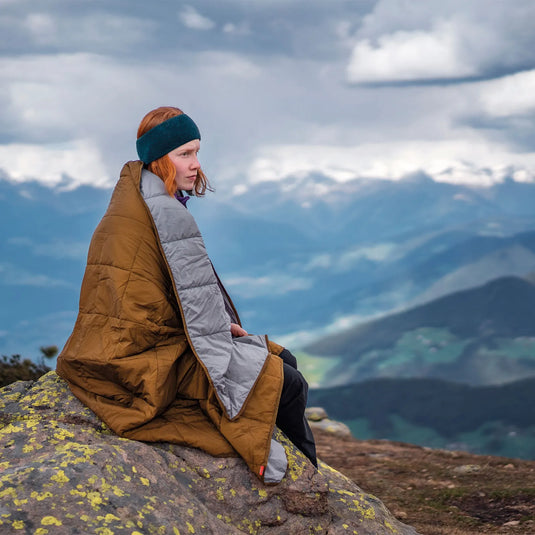 Eine Person sitzt auf einem Felsen vor Berglandschaft, eingehüllt in eine Decke.