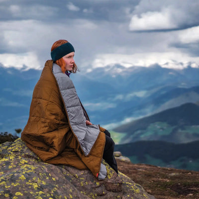 Eine in eine Robens® Icefall Quilt gehüllte Person sitzt auf einem Felsvorsprung mit Blick auf ferne Berge unter bewölktem Himmel.