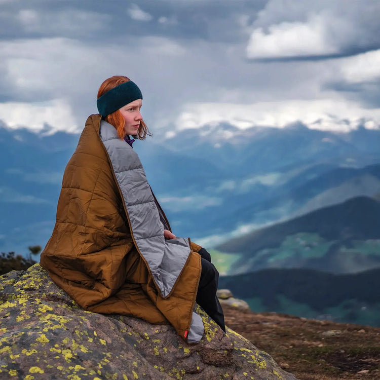 Eine in eine Robens® Icefall Quilt gehüllte Person sitzt auf einem Felsvorsprung mit Blick auf ferne Berge unter bewölktem Himmel.