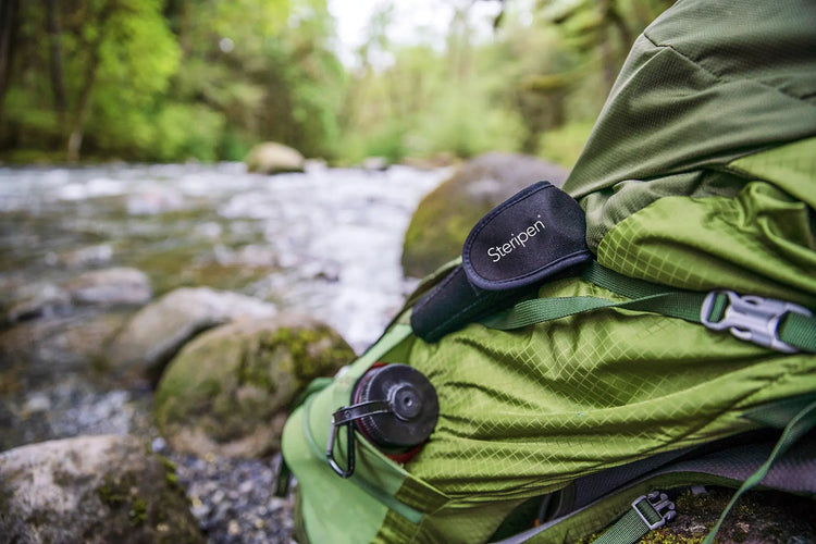 Ein grüner Rucksack, der auf einem Felsen an einem fließenden Bach in einem Waldgebiet ruht.