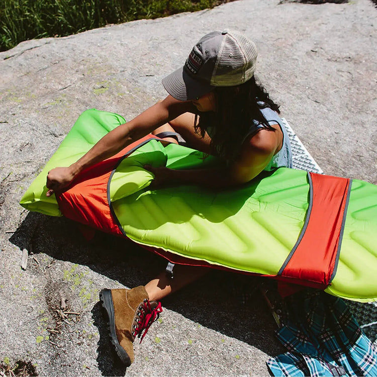 Frau sitzt auf einem Felsen mit einem Therm-a-Rest® Trekker Chair, trägt eine Mütze und Wanderstiefel und hält einen Outdoor-Ventilator.