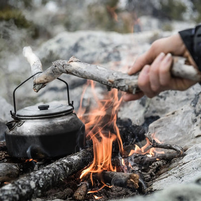 Eine Person legt Holz in ein Lagerfeuer, auf dem ein trangia® Wasserkessel steht.