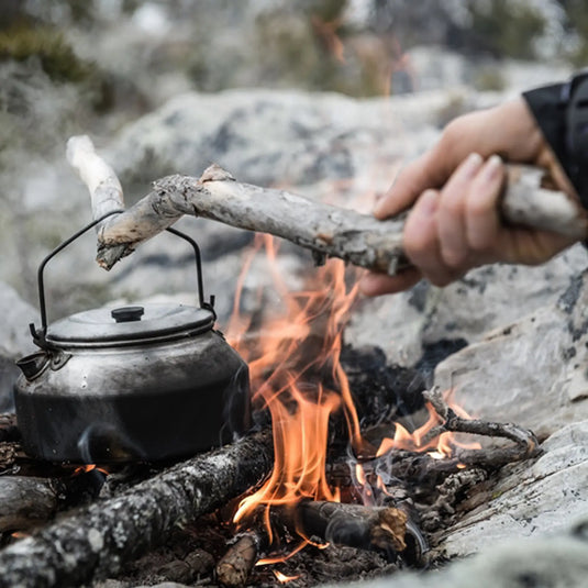 Eine Person legt Holz in ein Lagerfeuer, auf dem ein trangia® Wasserkessel steht.