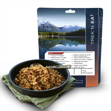 Abgepackte Outdoor-Mahlzeit von Trek'n Eat Jägertopf mit Rind und Nudeln neben einer schwarzen Schüssel mit dem zubereiteten Gericht, mit einem malerischen Bergsee-Hintergrund auf der Verpackung.