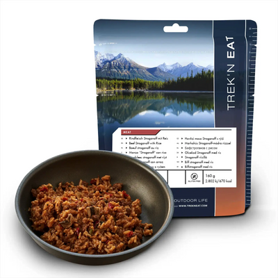 Eine Packung Trek'n Eat dehydriertes Rindfleisch Stroganoff mit Reis neben einer Schüssel mit der rehydrierten Mahlzeit, mit Naturhintergrund.