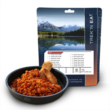 Eine Packung Trek'n Eat Chicken Tikka Masala dehydriertes Essen mit einer zubereiteten Mahlzeit, die in einer Schüssel daneben serviert wird, vor einem Hintergrund mit Bergen und einem See.