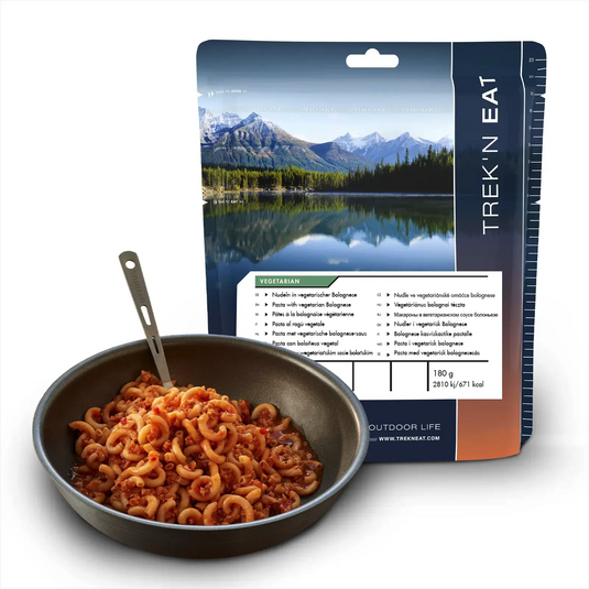 Eine Packung Trek'n Eat Nudeln in vegetarischer Bolognese neben einer Bratpfanne mit zubereitetem Nudelgericht.