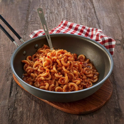 Eine Pfanne mit gekochten Trek'n Eat Nudeln mit vegetarischer Bolognese-Sauce auf einem Holztisch, dazu eine Gabel und eine karierte Serviette.