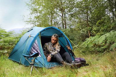 Eine Frau sitzt in einem blauen Vango™ „Alpha 250“ Kuppelzelt mit Campingausrüstung, umgeben von viel Grün.